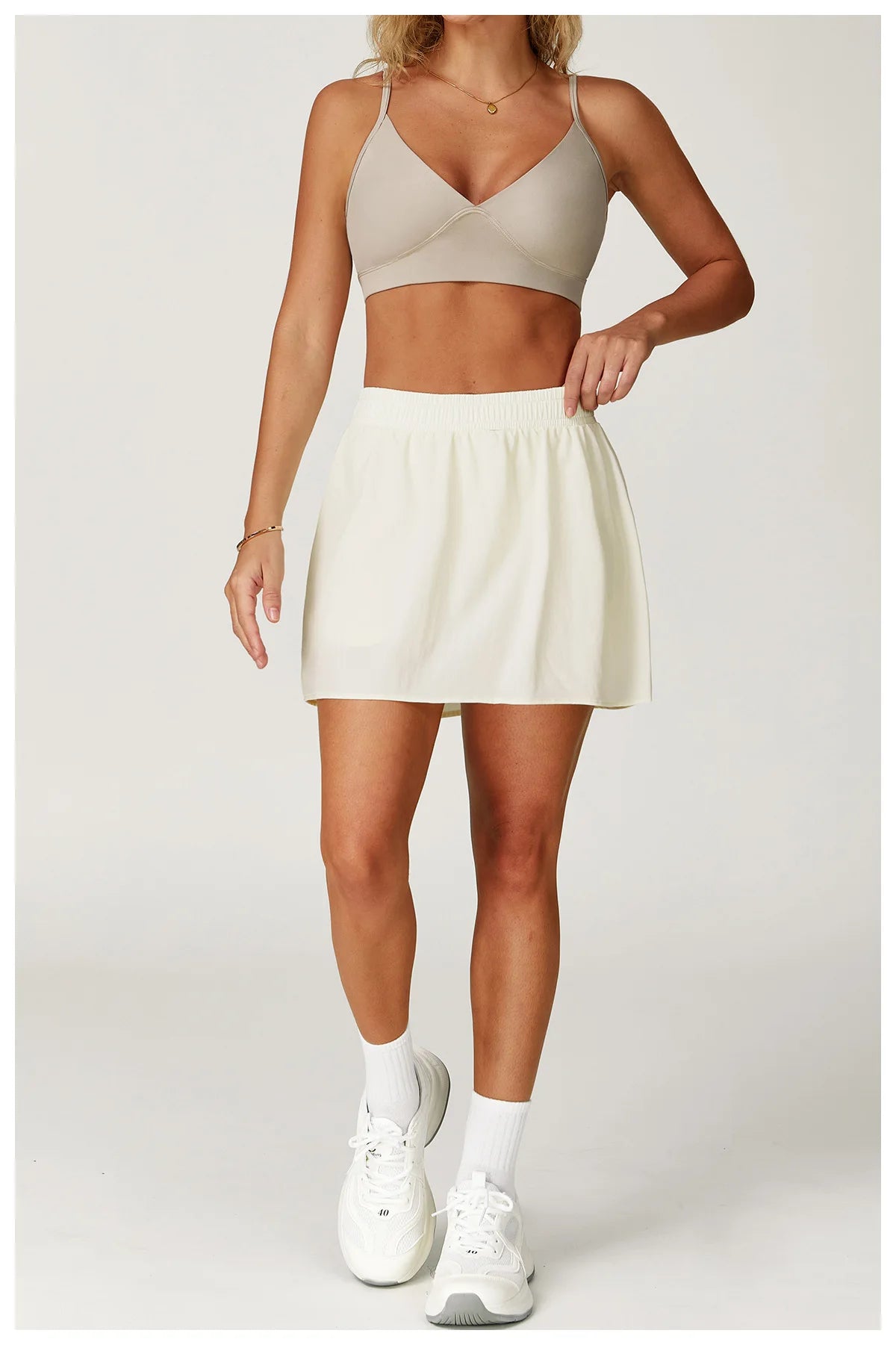 Woman modeling beige sports bra, white athletic skirt and sneakers on plain background
