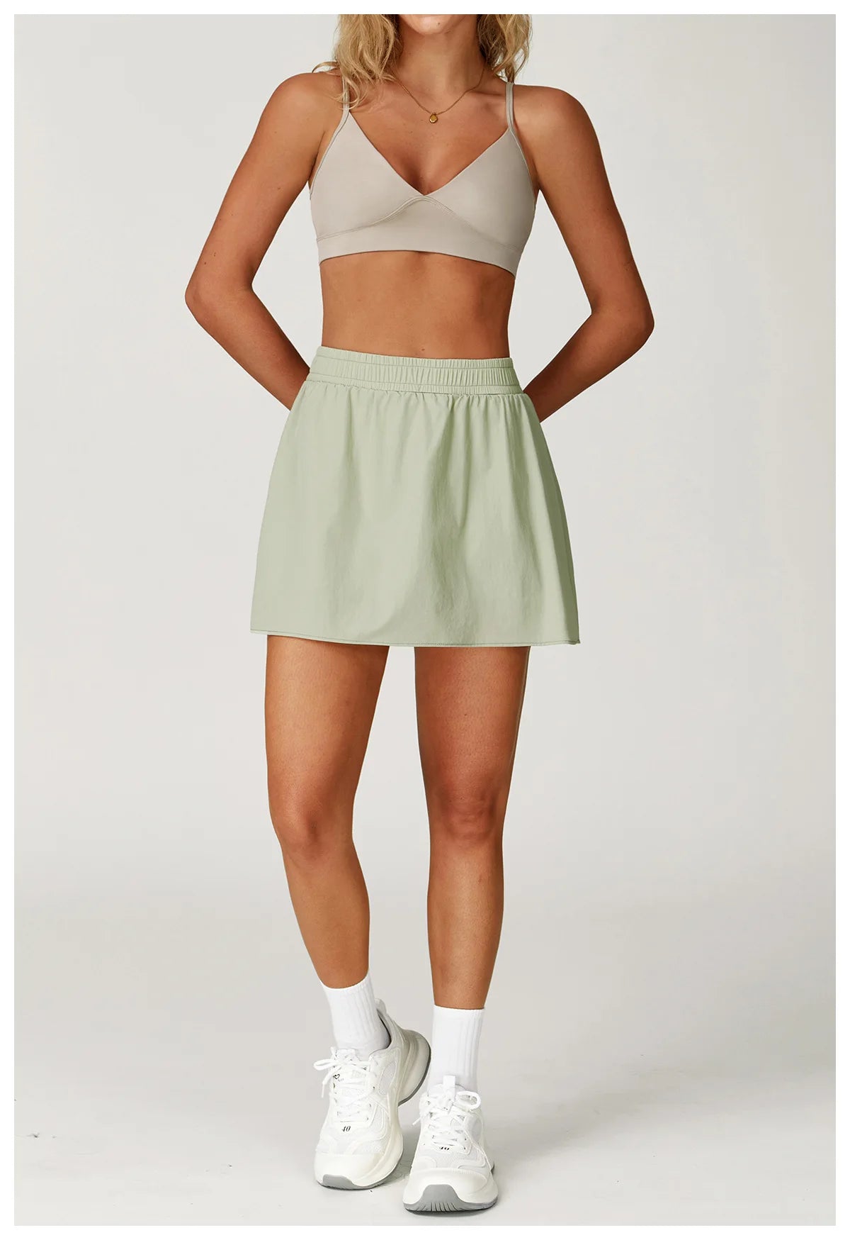 Woman in beige sports bra and light green athletic skirt, white sneakers, studio backdrop